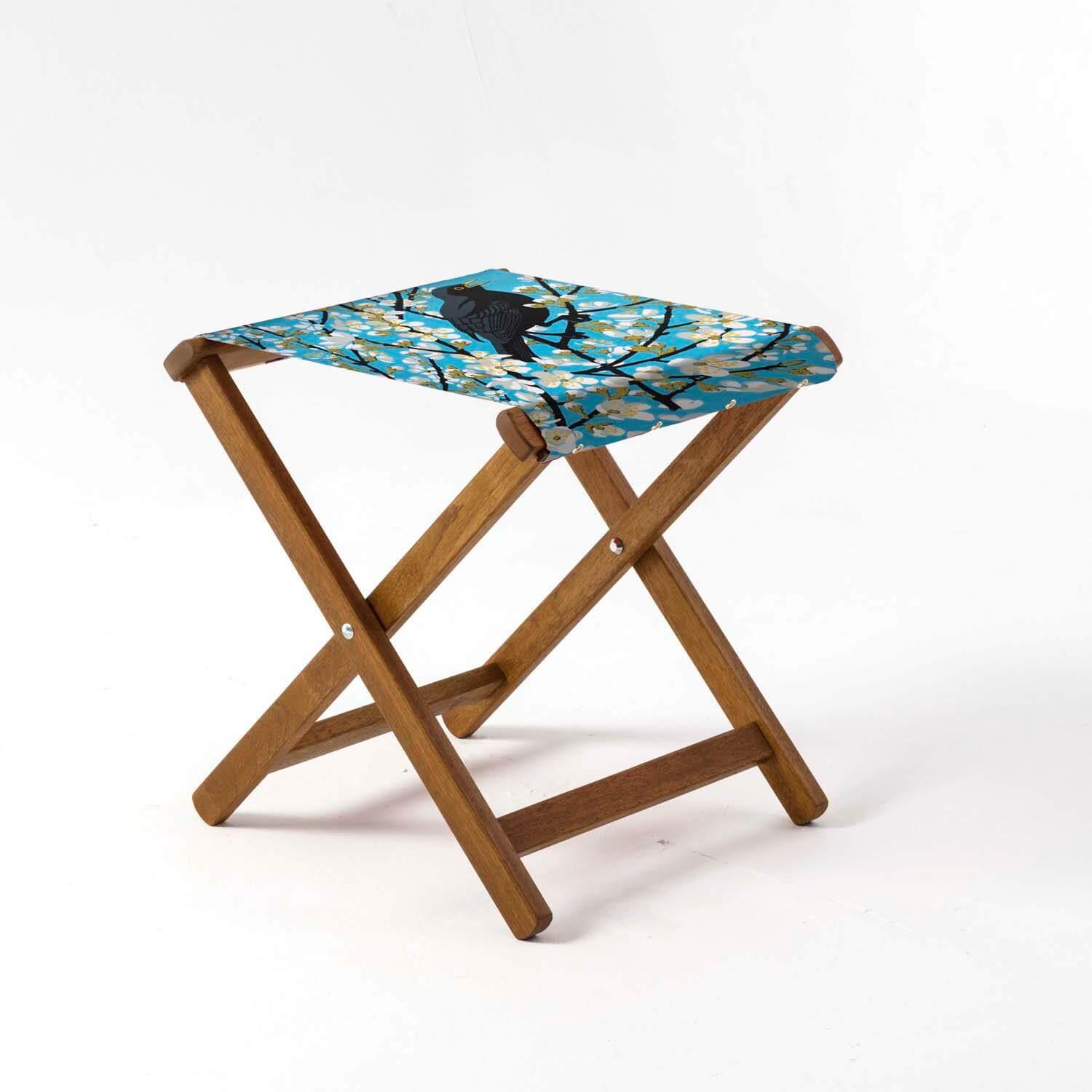 A wooden, folding stool. The fabric seat has a blue background a black bird on.