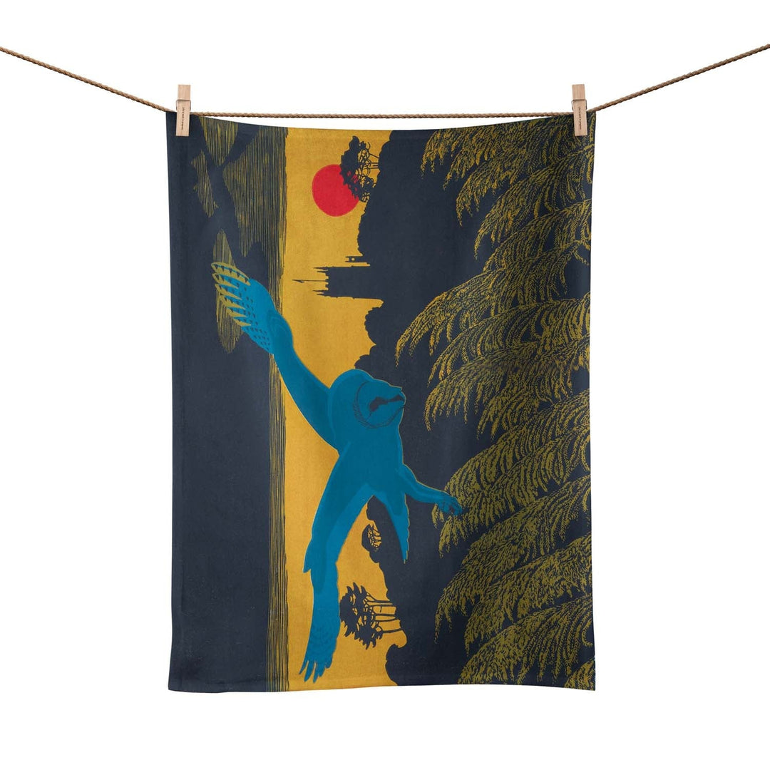 A tea towel pegged on a washing line. The tea towel shows a blue owl flying over a country scene.